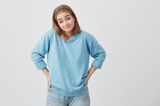 Clueless perplessa ragazza caucasica vestita in maglione e jeans scrollando le spalle, tenendo le mani sui fianchi e guardando con confusione, sopracciglia accigliate, non capendo cosa c'è che non va.