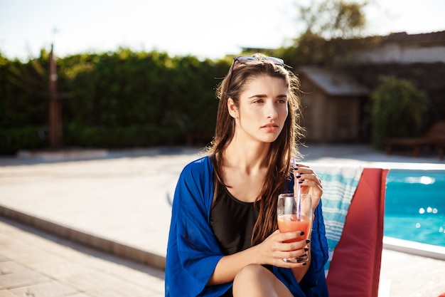 Cocktail bevente della bella donna castana, sedentesi vicino alla piscina