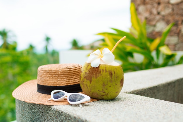 Cocktail di cocco decorato plumeria, cappello di paglia e occhiali da sole sul tavolo.