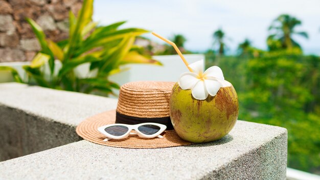 Cocktail di cocco decorato plumeria, cappello di paglia e occhiali da sole sul tavolo.