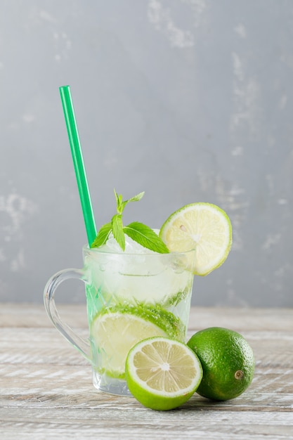 Cocktail ghiacciato di mojito in una tazza con calce, paglia, vista laterale della menta sul fondo di legno e gesso