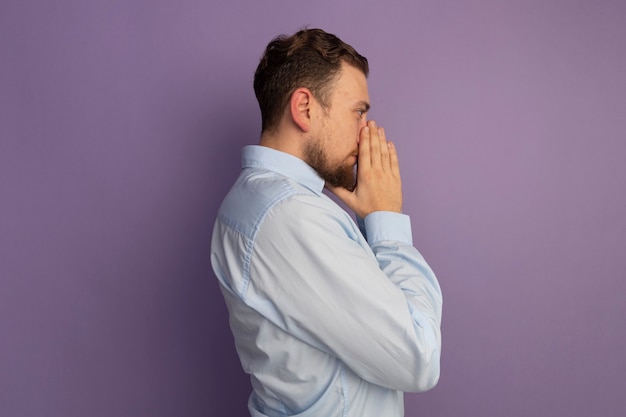 Cofident bell'uomo biondo sta lateralmente tenendo le mani insieme vicino al naso isolato sulla parete viola