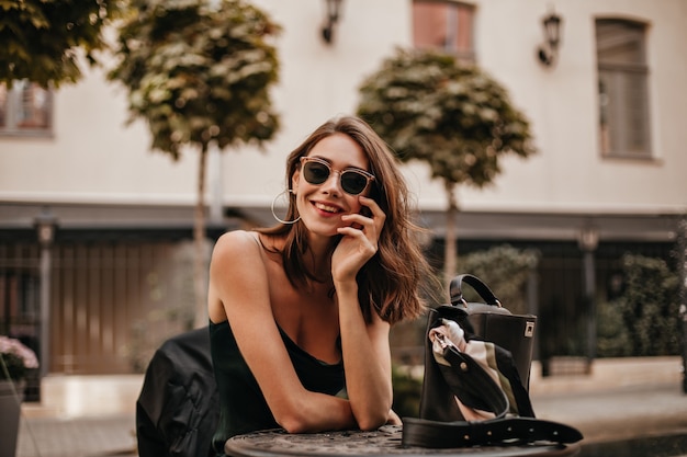 Coinvolgente giovane ragazza dai capelli scuri con labbra rosse, occhiali da sole moderni e abito sottoveste di seta, sorridente e in posa all'aperto