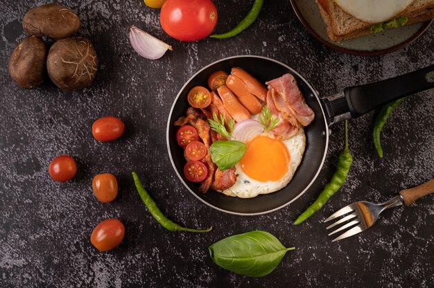 Colazione con uova fritte, salsiccia e prosciutto in padella con pomodori. Peperoncino e basilico