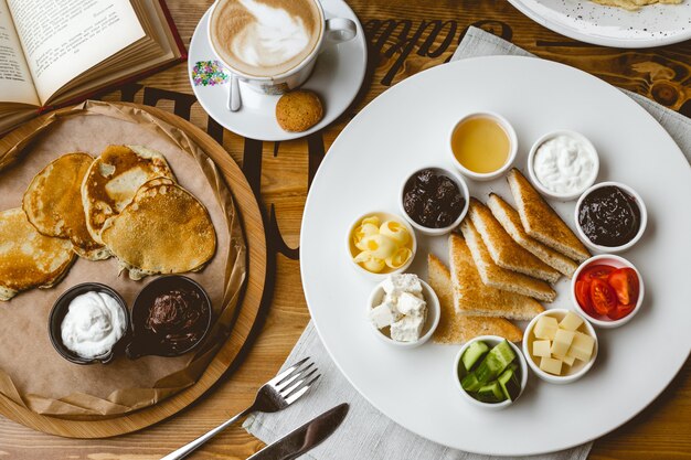 Colazione vista dall'alto set frittelle con crema al cioccolato e toast alla panna acida con marmellata crema al cioccolato miele cetriolo pomodoro burro e tazza di caffè sul tavolo