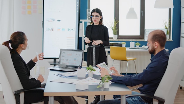 Colleghi che esaminano la presentazione in televisione con i grafici dei dati per lo sviluppo del business. Persone che lavorano per la pianificazione del progetto con laptop e tablet alla riunione aziendale. Compagni di lavoro con idee