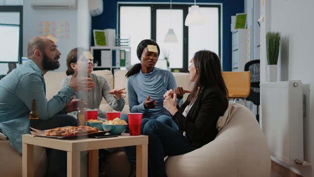 Colleghi che giocano a sciarade con note adesive dopo il lavoro in ufficio. Compagni di lavoro che si divertono a indovinare per fare attività divertenti e intrattenimento mentre bevono birra e mangiano pizza.