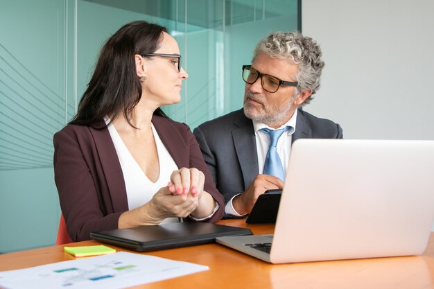 Colleghi che si incontrano e discutono del progetto, seduti al tavolo con il portatile aperto, utilizzando tablet e parlando.