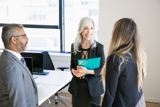 Colleghi di lavoro felici che indossano abiti, in piedi in ufficio, parlando e ridendo. Colpo medio. Concetto di colleghi