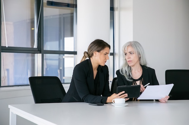 Colleghi femminili concentrati che discutono e analizzano i rapporti. Due professionisti seduti insieme, in possesso di documenti, utilizzando tablet e parlando. Concetto di lavoro di squadra