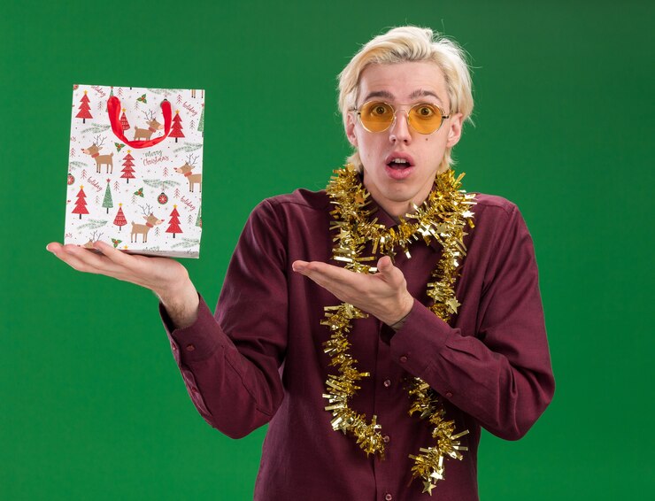 Foto gratuito colpito giovane uomo biondo con gli occhiali con ghirlanda di orpelli intorno al collo che tiene il sacchetto del regalo di natale che indica guardando la telecamera isolata su sfondo verde