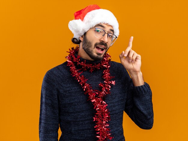 Colpito il giovane bel ragazzo che indossa il cappello di Natale con la ghirlanda sui punti del collo in alto isolato sulla parete arancione