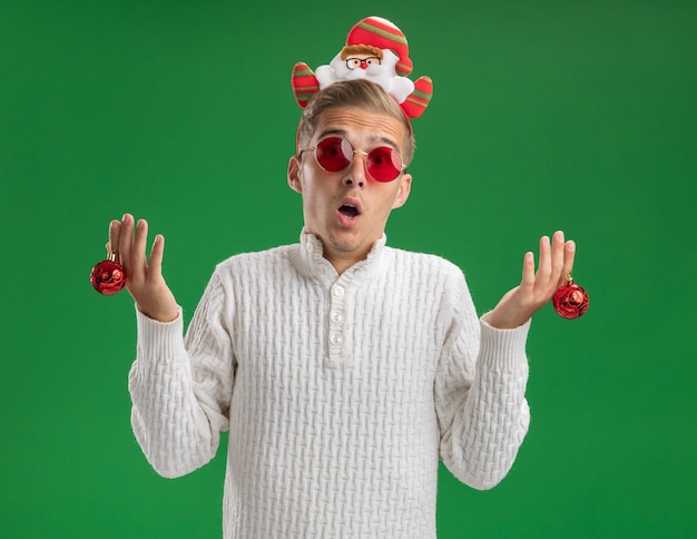 Colpito il giovane bel ragazzo che indossa la fascia di Babbo Natale con gli occhiali che tengono le palle dell'ornamento di Natale isolate sulla parete verde