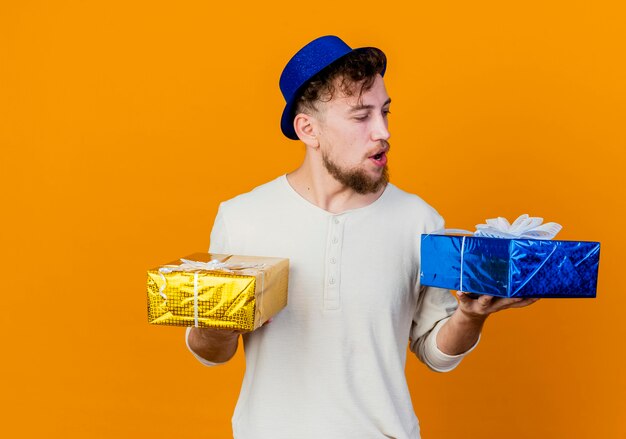 Colpito il giovane ragazzo slavo bello del partito che indossa il cappello del partito che tiene e che esamina le scatole regalo isolate su fondo arancio con lo spazio della copia