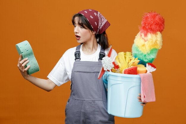 Colpito la giovane donna che indossa uniforme e bandana che tiene spugna e secchio di strumenti per la pulizia guardando la spugna isolata su sfondo arancione