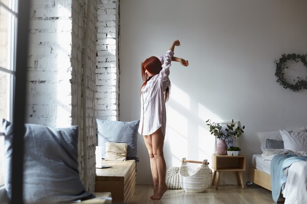 Colpo a figura intera di attraente giovane femmina rossa in camicia a righe lunghe in piedi a piedi nudi sulle dita dei piedi in camera da letto e allungando il corpo, alzando le braccia, di fronte alla grande finestra, godendosi la calda luce del sole primaverile