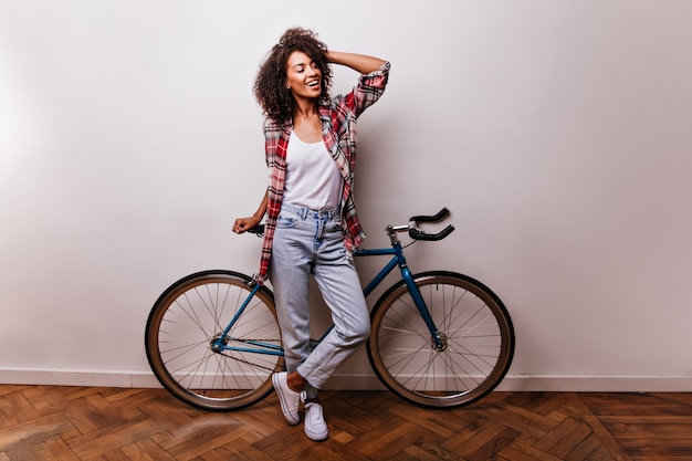 Colpo a figura intera di spettacolare signora riccia con la bicicletta. Ragazza nera ottimista che tocca i suoi capelli mentre posa.