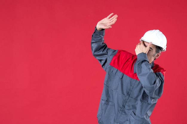 Colpo a metà corpo di un giovane architetto preoccupato emotivo in uniforme con elmetto e guardando in alto sul muro rosso isolato