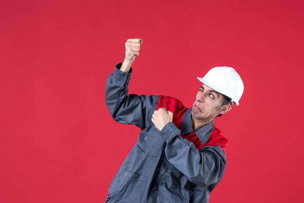 Colpo a metà corpo di un giovane costruttore emotivo divertente in uniforme con elmetto che mostra i suoi muscoli su un muro rosso isolato