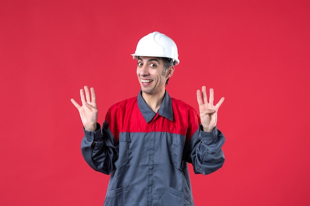 Colpo a metà corpo di un giovane costruttore sorridente in uniforme che indossa un elmetto che mostra quattro su un muro rosso isolato