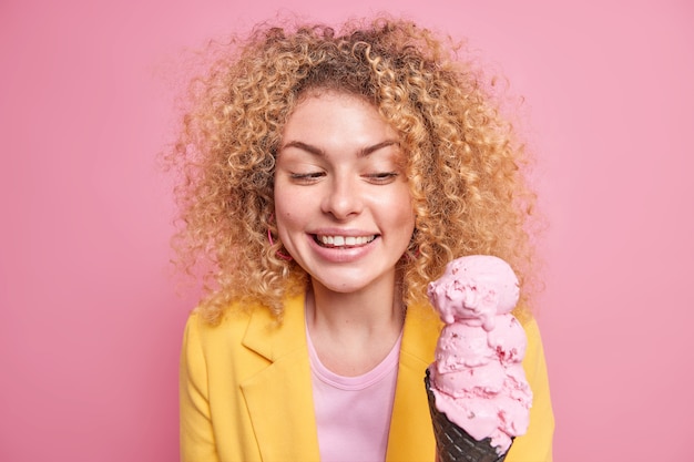 Colpo al coperto di bella donna europea allegra sorride felicemente guarda appetitoso gelato rosa in cialda nera indossa giacca gialla ha capelli ricci naturali isolati su muro roseo