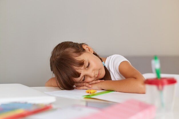 Colpo al coperto di piccola studentessa che dorme mentre è seduta al tavolo, stanca mentre fa i compiti, bambino con i capelli scuri che indossa una maglietta bianca.