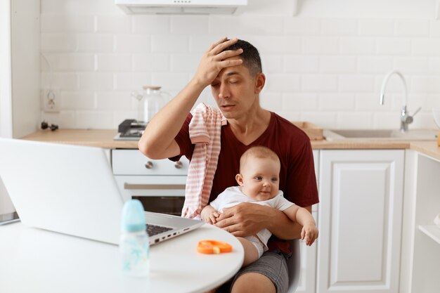 Colpo al coperto di un bell'uomo libero professionista stanco che indossa una maglietta bordeaux, in posa in cucina bianca, tenendo la mano sulla fronte, sente dolore alla testa, seduto davanti al computer portatile con il bambino in mano.