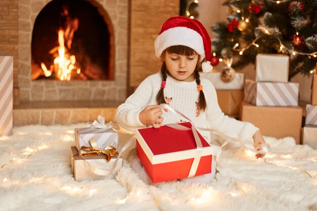 Colpo al coperto di una bambina carina che indossa un maglione bianco e un cappello di babbo natale, seduta sul pavimento vicino all'albero di Natale, scatole regalo e camino, con espressione concentrata durante l'apertura della confezione regalo.