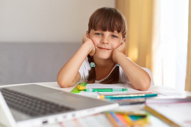 Colpo al coperto di una bambina felice positiva che guarda la telecamera con un'espressione facciale ottimista, facendo i compiti, ama le lezioni online durante la quarantena.