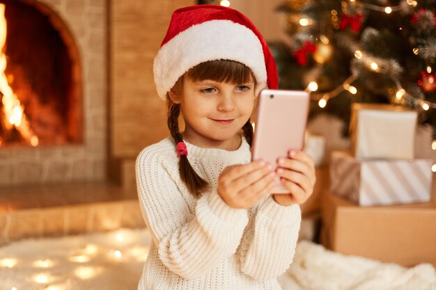 Colpo al coperto di una bambina sorridente felice che tiene in mano lo smartphone, indossa un maglione bianco e un cappello di babbo natale, seduta sul pavimento vicino all'albero di Natale, scatole regalo e camino.