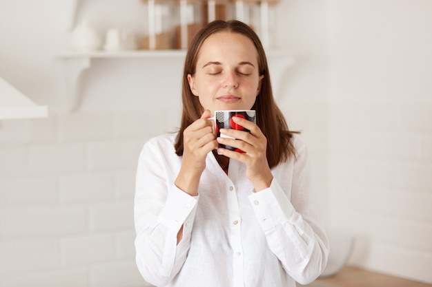 Colpo al coperto di una bella donna felice che si gode una tazza di caffè o tè in cucina al mattino, bevendo bevande calde, in piedi con gli occhi chiusi con espressione rilassata.