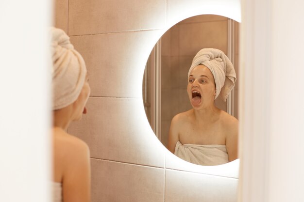 Colpo al coperto di una donna divertente in piedi in bagno con un asciugamano bianco sulla testa guardando il suo riflesso nello specchio con la bocca ampiamente aperta, mostrando la lingua fuori.
