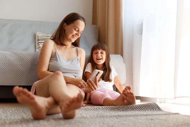 Colpo al coperto di una donna sorridente ottimista seduta sul pavimento vicino al divano e alla finestra con sua figlia carina e che tiene il telefono in mano, la famiglia ride felicemente.