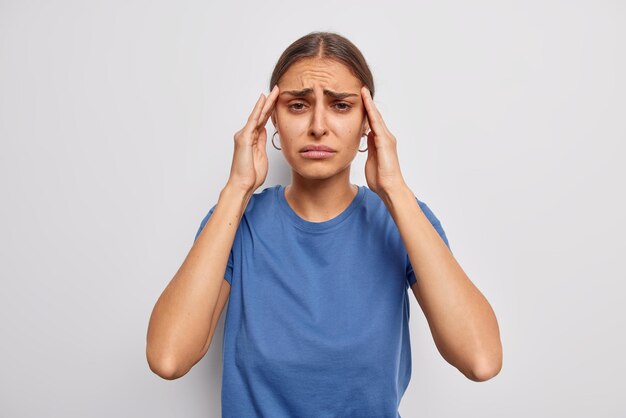 Colpo al coperto di una donna tesa infelice tiene le mani sulle tempie soffre di emicrania ha bisogno di antidolorifici indossa una maglietta blu casual isolata sul muro bianco