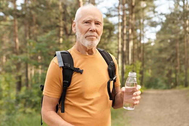 Colpo all'aperto del pensionato maschio fiducioso con la barba lunga con testa calva che tiene una bottiglia di acqua potabile, rinfrescandosi nella soleggiata giornata estiva mentre si viaggia da soli nella natura selvaggia con lo zaino