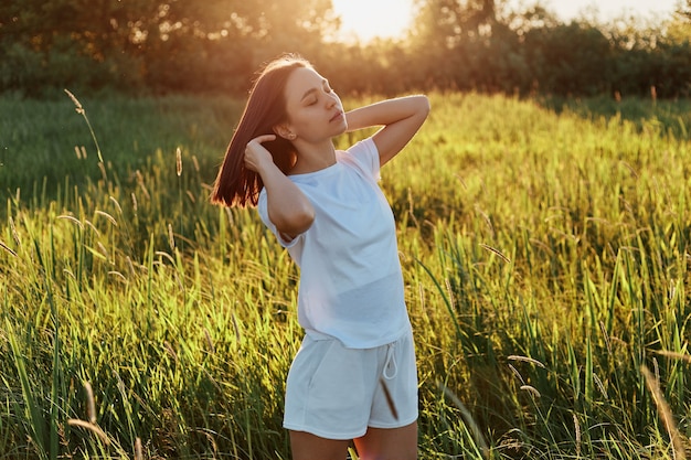 Colpo all'aperto di attraente donna dai capelli scuri che indossa una maglietta bianca e distoglie lo sguardo, alzando le braccia, posando nel prato verde, godendosi il bellissimo tramonto e la natura.