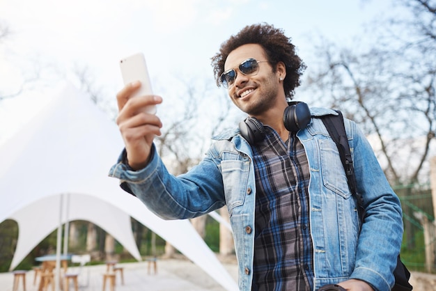 Colpo all'aperto di attraente giovane afroamericano con acconciatura afro e cuffie sul collo che indossa abiti alla moda e occhiali che tengono smartphone mentre registrano la band di strada nel parco