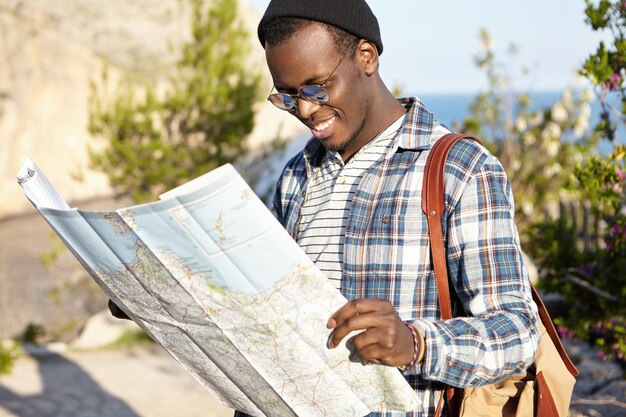 Colpo all'aperto di felice sorridente giovane attraente turista africano nel pittoresco paesaggio leggendo la mappa cartacea, alla ricerca di percorsi e nuove visite turistiche, indossando occhiali da sole rotondi alla moda con lenti a specchio