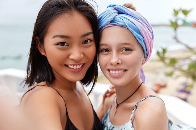 Colpo all'aperto di turisti femminili felici che passeggiano vicino al mare, respirano fresca brezza marina, fanno selfie, hanno sorrisi positivi sui volti. La femmina europea con la sciarpa sulla testa incontra la sua amica asiatica