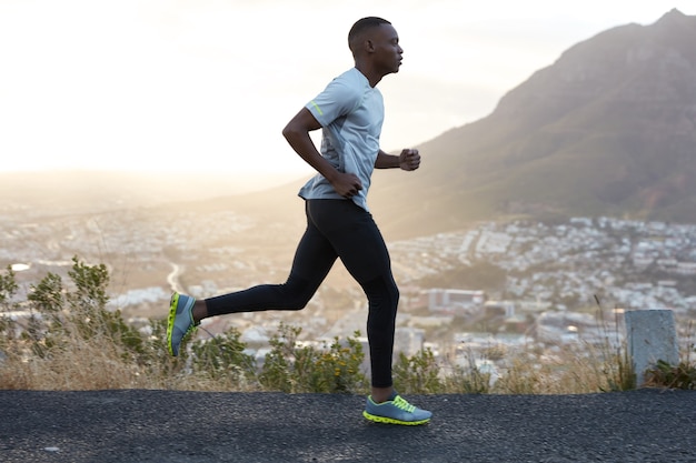 Colpo all'aperto di un uomo attivo dalla pelle scura che corre al mattino, ha allenamenti regolari, vestito con tuta da ginnastica e scarpe da ginnastica comode, concentrato nella distanza, vede finire lontano.