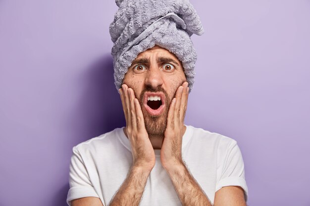 Colpo alla testa di un bell'uomo che tocca le guance, ha un'espressione del viso insoddisfatta, si rende conto di avere una pelle problematica, tiene le mani sulle guance, indossa una maglietta bianca casual, un asciugamano avvolto sulla testa, sta al coperto