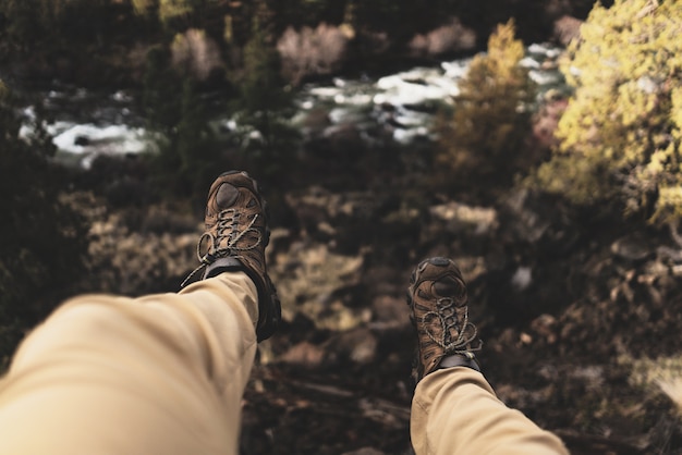 Colpo ambientale selettivo di una persona che indossa le scarpe marroni di sport d'escursione che si siedono su una scogliera vicino agli alberi