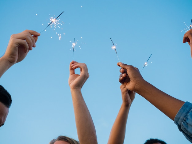 Colpo del primo piano dei giovani con le luci del Bengala in mani sollevate. Gruppo di amici che si diverte alla festa all'aperto. Concetto di celebrazione