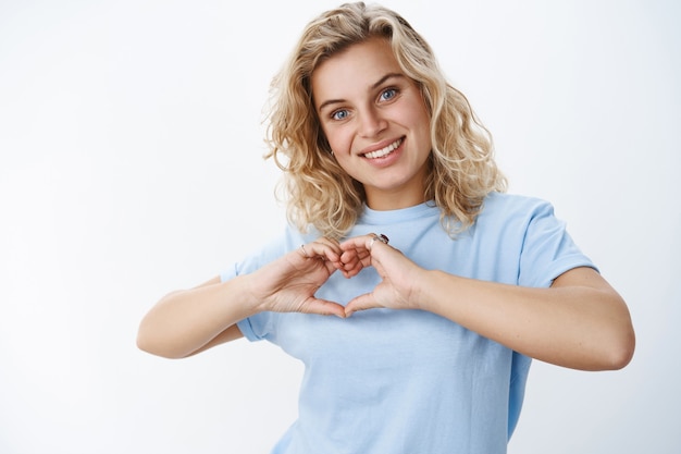 Colpo del primo piano della ragazza romantica e carina con gli occhi azzurri e taglio di capelli biondo corto sorridente caldo e dolce, mostrando il segno del cuore sul petto esprimendo amore e simpatia, facendo un gesto commovente