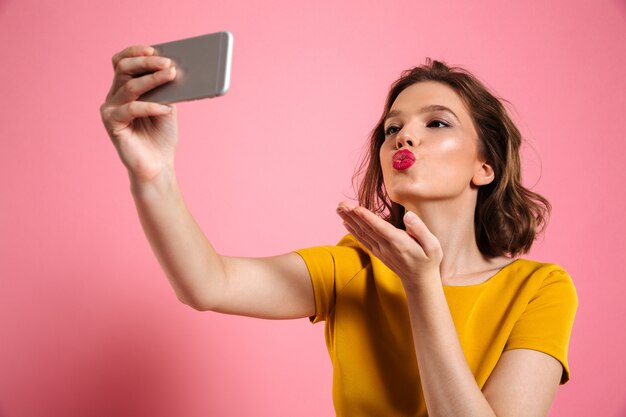 Colpo del primo piano di giovane donna attraente con trucco luminoso che invia bacio dell'aria mentre prendendo selfie sul telefono cellulare