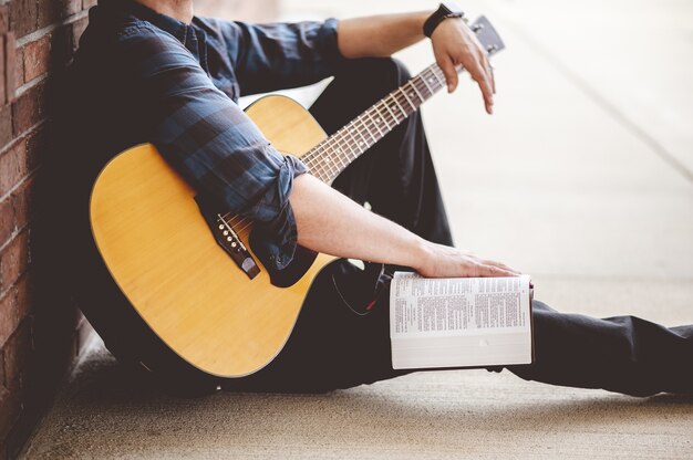 Colpo del primo piano di un giovane maschio seduto con un libro e una chitarra in mano