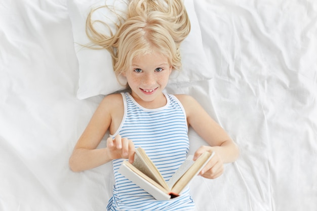 Colpo dell'interno della bambina adorabile allegra con capelli biondi che si trovano sul cuscino bianco nella sua camera da letto, godendo leggendo favola. Carino bambina dagli occhi azzurri che legge invece di fare un sonnellino, con uno sguardo furbo