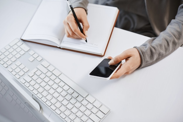 Colpo di alto angolo di mani di donna che lavorano con gadget. Colpo potato dello smartphone femminile moderno della tenuta mentre scrivendo piano in taccuino, sedendosi vicino alla tastiera e al computer, avendo tempo difficile all'ufficio