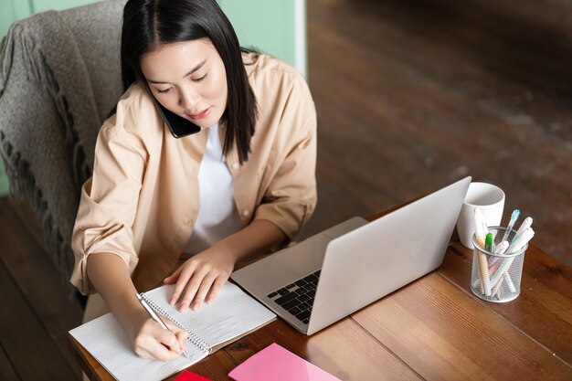 Colpo di angolo superiore della donna asiatica che studia sul telefono che parla sul telefono cellulare e che annota le note wor...