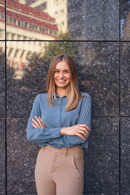 Colpo di bella giovane donna di affari che indossa la camicia di chiffon blu mentre levandosi in piedi con le braccia conserte sul muro di marmo grigio